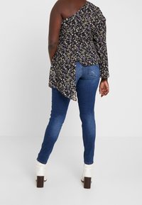 Top floral negro asimétrico con una sola manga larga, combinado con jeans azules y botas de tobillo blancas con tacón. Material texturizado.