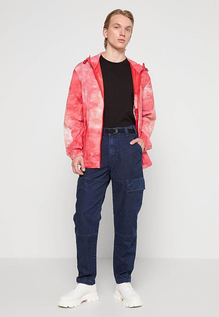 Jonge man staat in een rood tie-dye capuchonjack, zwarte shirt, donkerblauwe cargobroek en witte schoenen met dikke zool.