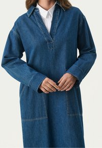 Denim-paitapusero, jossa on syvä V-kaula-aukko, pitkät hihat ja kaksi etutaskua. Omistaa sinisen pesun ja erottuvat ompeluyksityiskohdat.