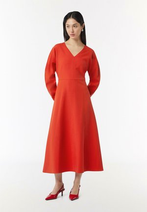 Rotes langärmliges Kleid mit V-Ausschnitt und taillierter Oberseite, das zu einem weiten Rock ausläuft. Kombiniert mit glänzenden roten High Heels.