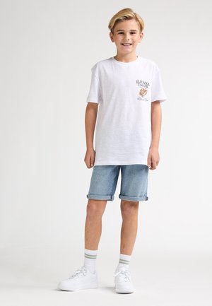 T-shirt blanc à manches courtes avec impression graphique, associé à un short en denim bleu clair retroussé et des baskets blanches avec des accents gris.