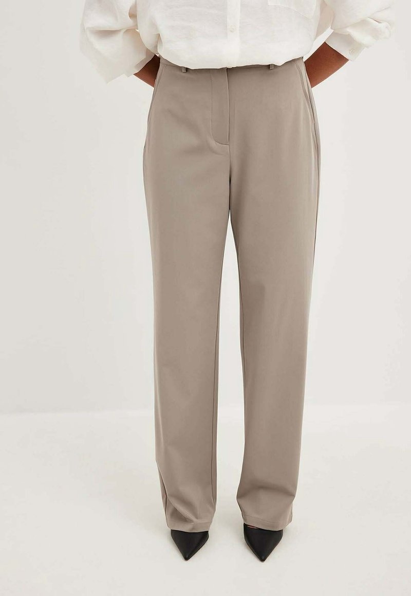 Personne portant un pantalon droit beige clair, une chemise blanche aux manches retroussées et des chaussures noires pointues, les mains dans les poches, debout à l'intérieur.