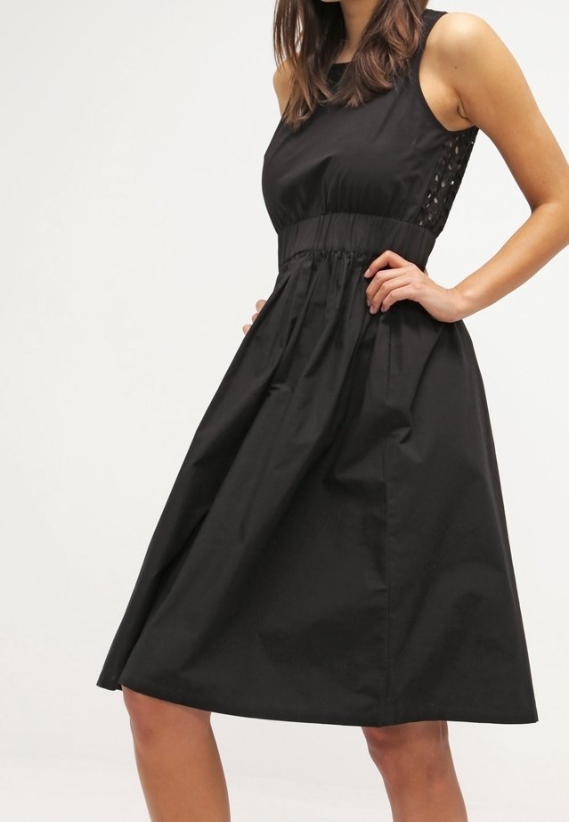 Femme portant une robe midi noire sans manches avec taille cintrée et découpes latérales, debout les mains sur les hanches devant un fond blanc.