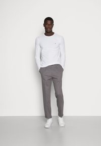 Langærmet hvid skjorte med et lille logo på brystet, parret med grå skræddersyede bukser og hvide sneakers, stående mod en neutral baggrund.