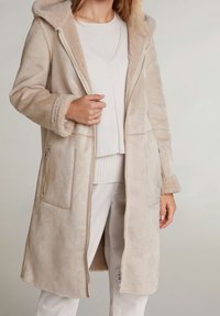 Kvinne iført en beige lang shearlingjakke med glidelåslommer og mansjetter, kombinert med hvit genser og hvite bukser.