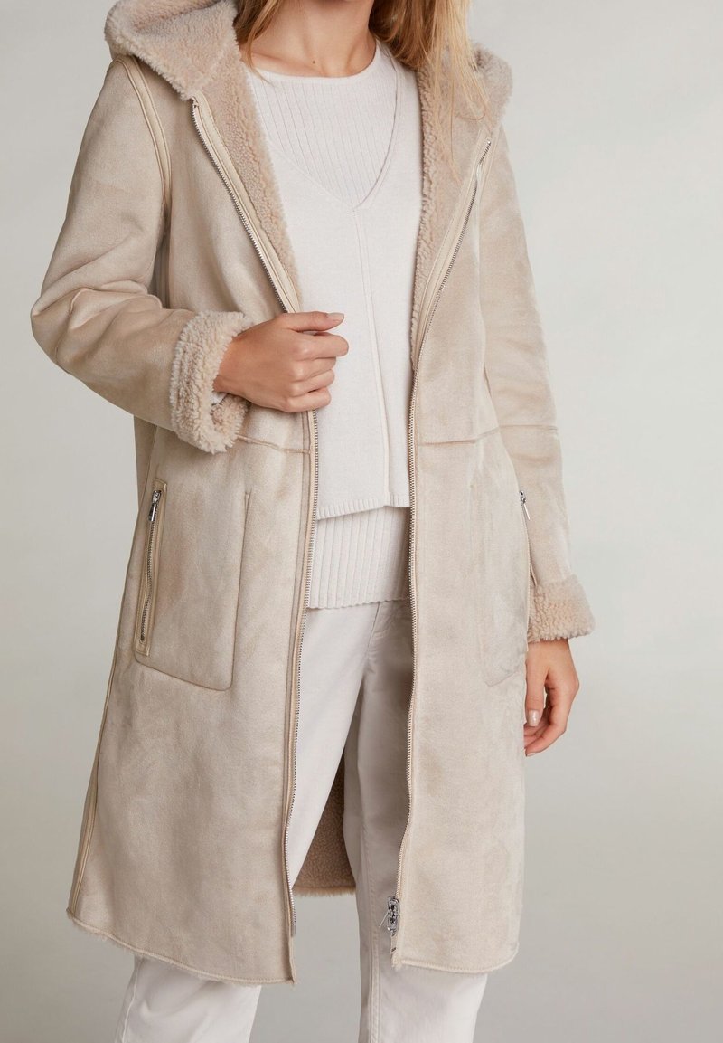 Kvinne iført en beige lang shearlingjakke med glidelåslommer og mansjetter, kombinert med hvit genser og hvite bukser.