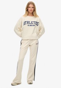 Sudadera color crema con el texto "ATHLETICS" en azul marino, combinada con pantalones acampanados color crema que tienen franjas laterales oscuras y zapatillas casuales.