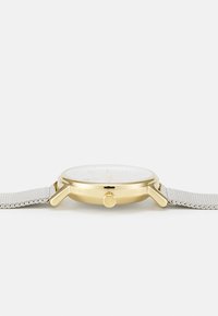 Kulaté hodinky se zlatým nerezovým pouzdrem, bílým ciferníkem, stříbrným síťovým páskem a postranní korunkou pro nastavení.