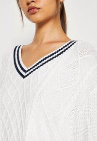 Vit stickad tröja med ett texturerat diamantmönster och en djup V-ringning med marinblå randig kant.