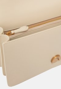 Borsa a mano in pelle beige con chiusura zip dorata, texture liscia e accento con logo rotondo. Linee pulite e design minimalista.