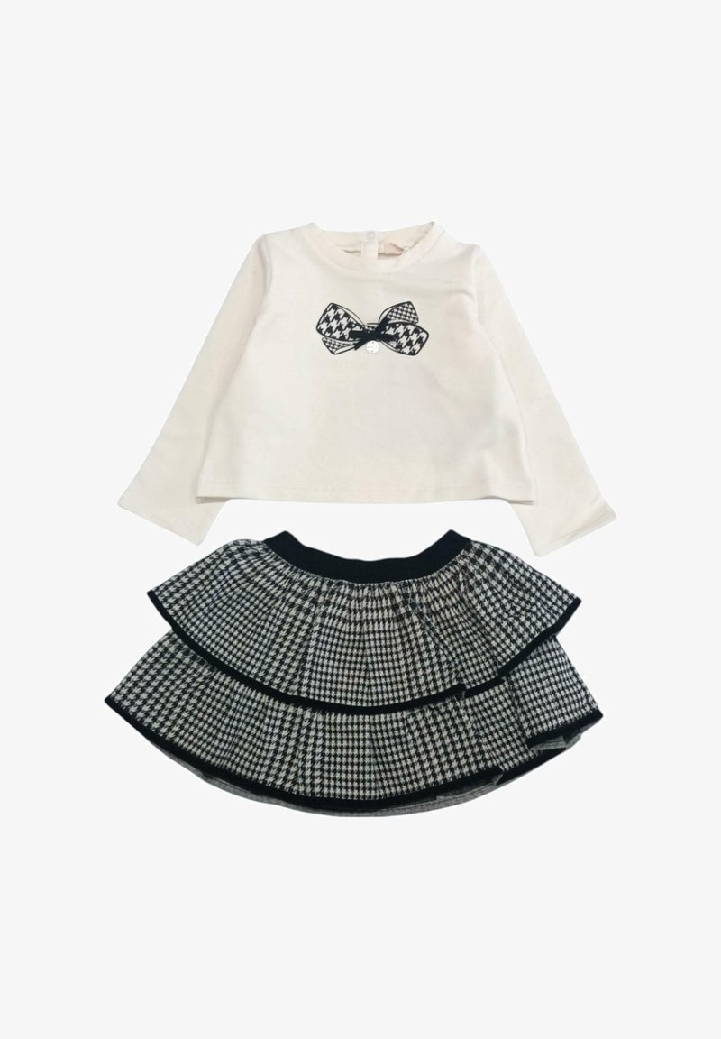 Weißes Langarmoberteil mit schwarzem Schleifenmotiv, kombiniert mit einem schwarz-weißen Hahnentrittmuster-Rock mit mehreren Lagen und elastischem Bund.