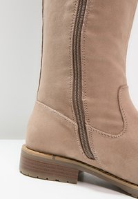 Bota de ante beige hasta la rodilla con textura suave, cremallera lateral y un tacón de madera apilada para mayor estabilidad.