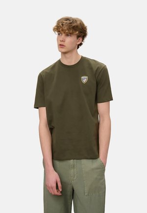 Giovane uomo con capelli ricci che indossa una maglietta verde oliva con un piccolo logo sul petto e pantaloni verde chiaro, con le mani in tasca.