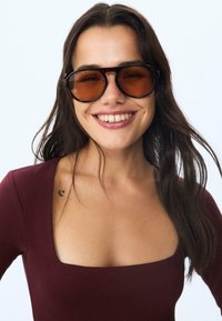 Sonnenbrille mit großem quadratischem Rahmen in Dunkelbraun, ausgestattet mit bernsteinfarbigen Gläsern. Das Modell trägt ein figurbetontes bordeauxfarbenes Top mit quadratischem Ausschnitt.
