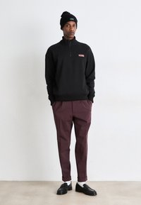 Μαύρο φούτερ με φερμουάρ και γιακά, που έχει κόκκινο λογότυπο, συνδυασμένο με μπορντό κομμένα παντελόνια και μαύρες loafers με λευκές κάλτσες.