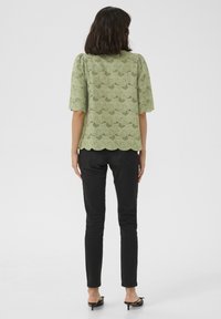 Top verde chiaro con orlo a onde e design in pizzo floreale, maniche corte, abbinato a pantaloni skinny neri e sandali con tacco neri.