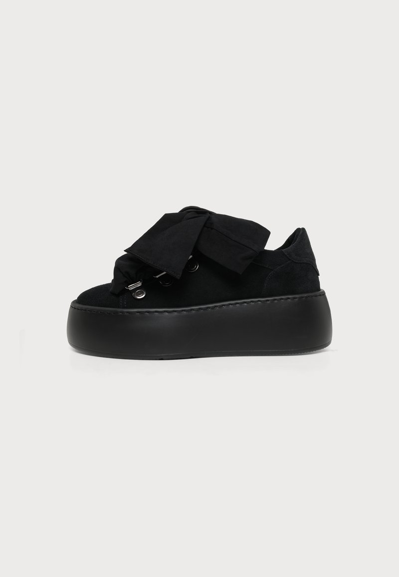 Μαύρο platform sneaker με παχιά σόλα, επάνω μέρος από σουέτ, μεταλλικά μεταλλικά ακροδέκτες και φαρδιά δεμένα υφασμάτινα κορδόνια σε απλό φωτεινό φόντο.