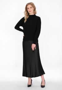 Schwarzes Langarmoberteil mit Stehkragen, kombiniert mit einem fließenden schwarzen Satin-Maxikleid und schwarzen High Heels. Glatte Textur, minimalistisches Design.