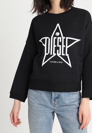 Czarna bluza z długim rękawem z dużą białą gwiazdą i napisem "DIESEL", w parze z jasnoniebieskimi dżinsami.