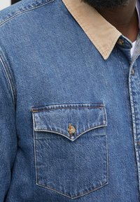 Denimskjorta med krage i beige kordflors, framficka och kontrasterande sömnadsdetaljer. Den blåa tyget har en texturerad yta.