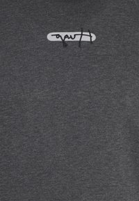 T-shirt in cotone grigio con un breve logo ricamato bianco, posizionato leggermente decentrato sul lato sinistro superiore.