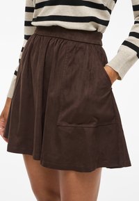 Rjava semiš krilo na zvonec z elastičnim pasom in stranskimi žepi. Gladek tekstura, dolžina do kolen. Delno viden črtast zgornji del.