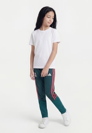 Fille debout, portant un t-shirt blanc uni, un pantalon de survêtement Adidas vert avec des bandes rouges, et des baskets blanches, regardant vers le bas à sa gauche.