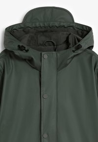 Chaqueta de lluvia verde impermeable con capucha, que cuenta con botones a presión, un forro interior texturizado y cordones ajustables en el cuello.