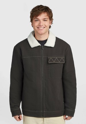 Junger Mann lächelt, trägt eine dunkle Reißverschlussjacke mit weißem Fleece-Kragen und aufgenähter Taschenklappe, steht vor einem einfarbigen hellen Hintergrund.