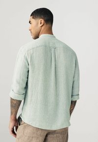 Camisa de manga larga de color verde claro y texturizada, con un diseño de manga enrollable y un pliegue en la parte posterior. La tela parece transpirable y casual.