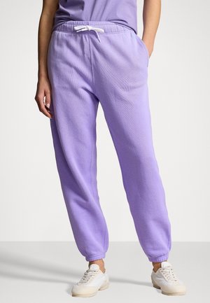 Persona che indossa pantaloni della tuta color lilla chiaro con coulisse bianca e scarpe da ginnastica bianche, in piedi con una mano in tasca su sfondo semplice.