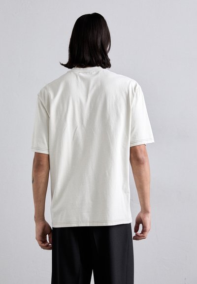 Világosszürke, rövid ujjú pamut póló kerek nyakkal, laza szabással és finom varrási részletekkel az alsó szegélyeken. A hátulnézete látható.