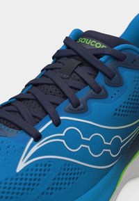 Μπλε αθλητικό παπούτσι με μαύρα κορδόνια, λευκό λογότυπο στο πλάι, πράσινο κείμενο στη μαύρη γλώσσα και διαπνέον δίχτυ ύφασμα.