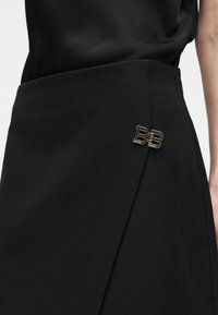 Jupe portefeuille noire avec une broche logo "BB" en ton doré sur le côté, associée à un haut noir, montrant une partie du bras et de la taille d'une personne.