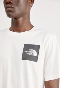 Bijela pamučna majica s crnim kvadratnim logom s natpisom "THE NORTH FACE" i zakrivljenim dizajnom, okrugli izrez, kratki rukavi.