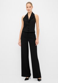 Schwarzes ärmelloses Oberteil mit Punkten, einem eingekerbten Kragen und Knöpfen vorne, kombiniert mit schwarzen Weitbein-Hosen und High-Heels.