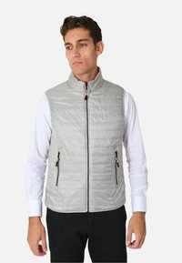 Gilet imbottito grigio chiaro con colletto alto, due tasche con zip e dettagli arancioni sulla zip. Indossato sopra una camicia bianca.