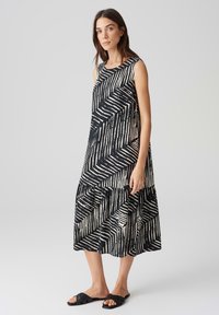 Abito senza maniche bianco e nero con un motivo geometrico a zigzag, silhouette a lineafluida e orlo a strati, indossato con sandali neri piatti.