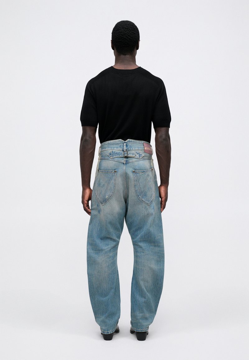 Svart kortärmad skjorta och ljusblå vida jeans med en urtvättad textur och logotypdetalj på midjebandet, sedd bakifrån.