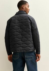 Pikowana ciemnoszara kurtka, z kołnierzem, kieszeniami bocznymi i diamentowym wzorem. Połączona z niebieskimi dżinsami. Widok z tyłu.