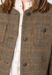 Bruine tweedjas met visgraatpatroon, voorzien van twee borstzakken en gouden knopen, gecombineerd met een witte blouse met kraag.