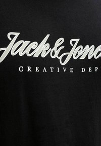 Logo Jack & Jones en cursive blanche avec « CREATIVE DEP » en majuscules, police serif, sur fond en tissu noir.