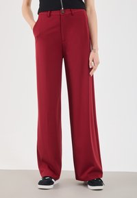Red wide-leg trousers made of smooth fabric with a button closure and side pockets, paired with black slip-on sneakers. Plain background.
