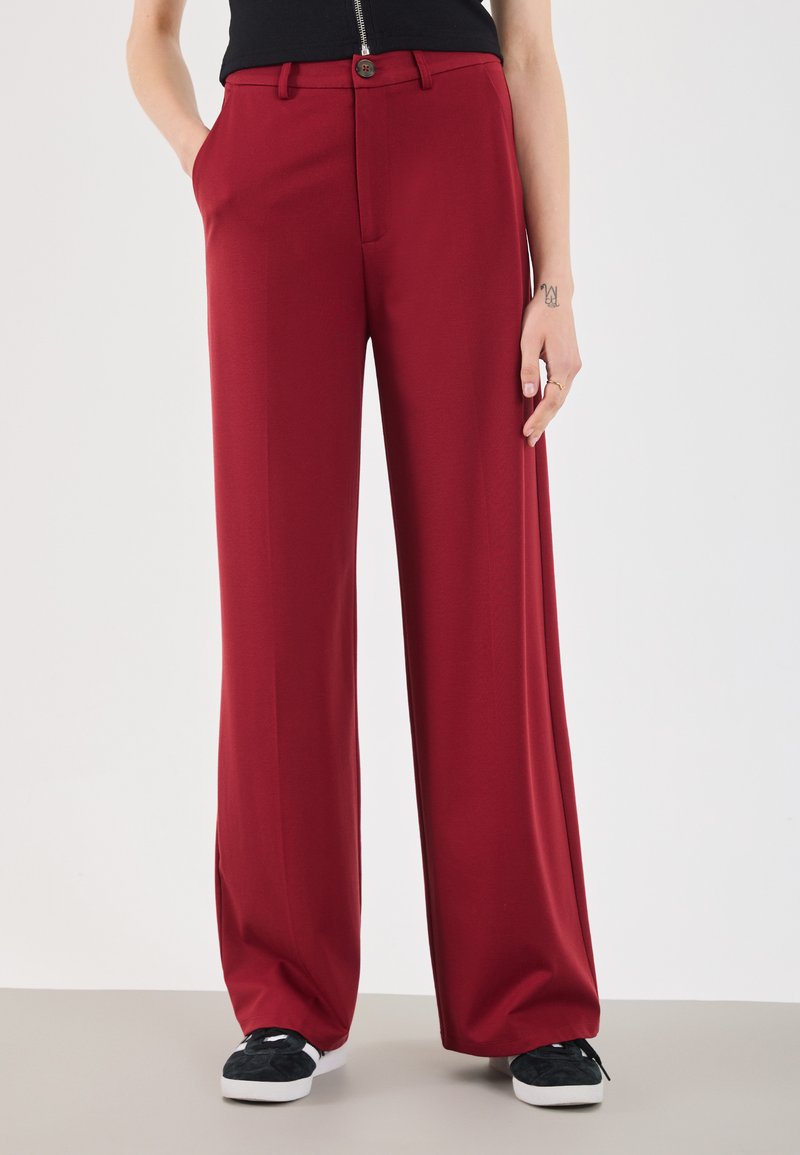 Red wide-leg trousers made of smooth fabric with a button closure and side pockets, paired with black slip-on sneakers. Plain background.