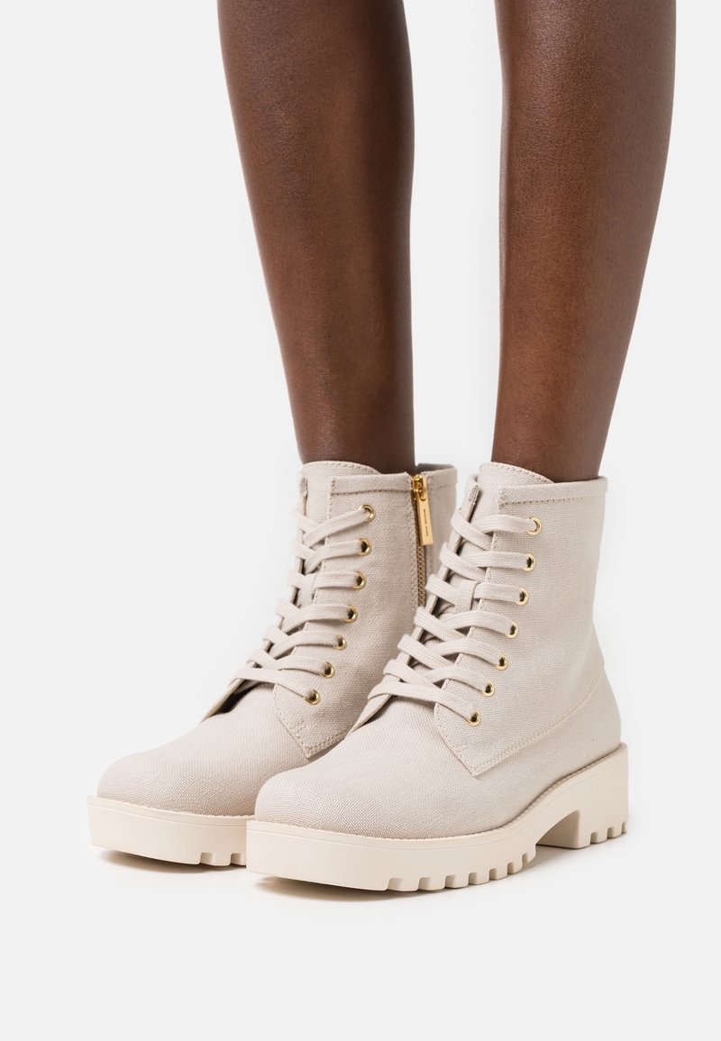Gros plan sur des jambes à la peau foncée portant des bottines beige à lacets, avec des semelles épaisses et une fermeture éclair dorée sur le côté, sur fond blanc.