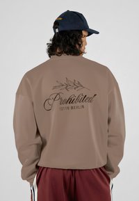 Brun oversized genser med en glatt tekstur. Har "Forbudt" og "10119 BERLIN" trykket i mørkebrun på baksiden, over et bladdesign.