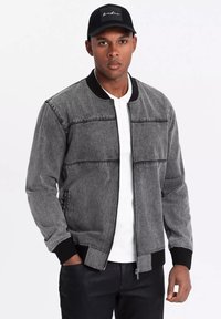 Giubbotto bomber in denim grigio con colletto e polsini a coste neri, caratterizzato da cuciture orizzontali. Indossato sopra una camicia bianca, con un cappellino nero.