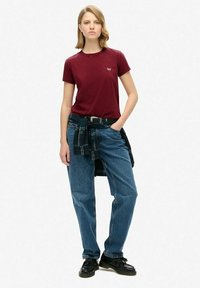 Vrouw in een bordeauxrode t-shirt, blauwe jeans, zwarte schoenen en een geruit overhemd om haar taille geknoopt, staande met één hand in haar zak.