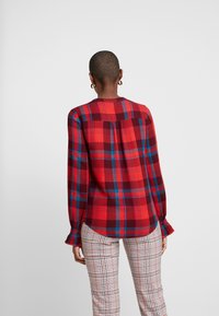 Chemise à carreaux rouges et bleus avec des manches longues, des poignets froncés et un col rond, associée à un pantalon à motifs à carreaux.