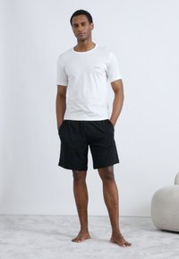 Camiseta blanca de cuello redondo con un pequeño logo rojo, combinada con pantalones cortos negros. El modelo está de pie de manera casual sobre una superficie alfombrada clara.